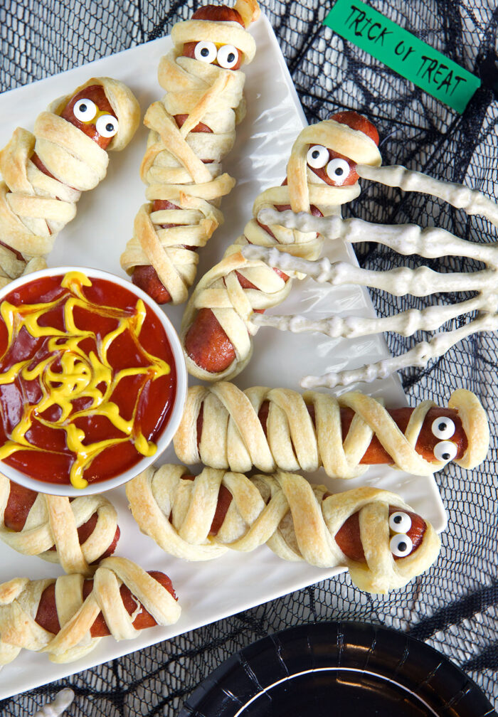 Mummy hot dogs are placed on a white plate with ketchup and mustard in the middle.
