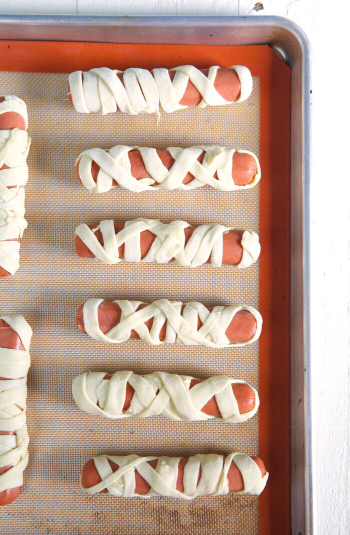 Puff pastry wrapped hot dogs are lined up on a baking sheet, ready to go into the oven.
