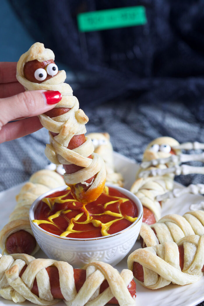A mummy hot dog is being dipped into ketchup.
