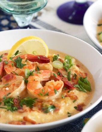 White bowl with shrimp and grits with a slice of lemon and a wine glass in the background.