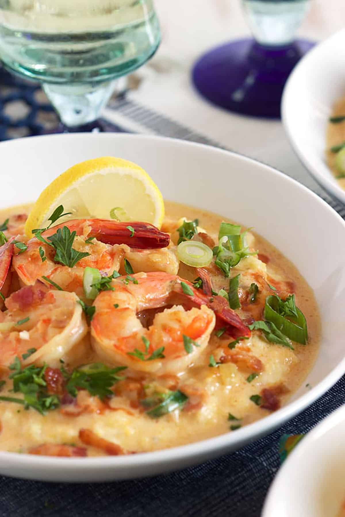 White bowl with shrimp and grits with a slice of lemon and a wine glass in the background.