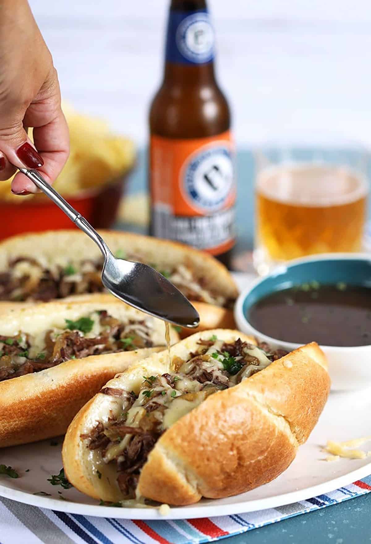 Three French Dip Sandwiches with a spoon drizzling au jus on a sandwich.