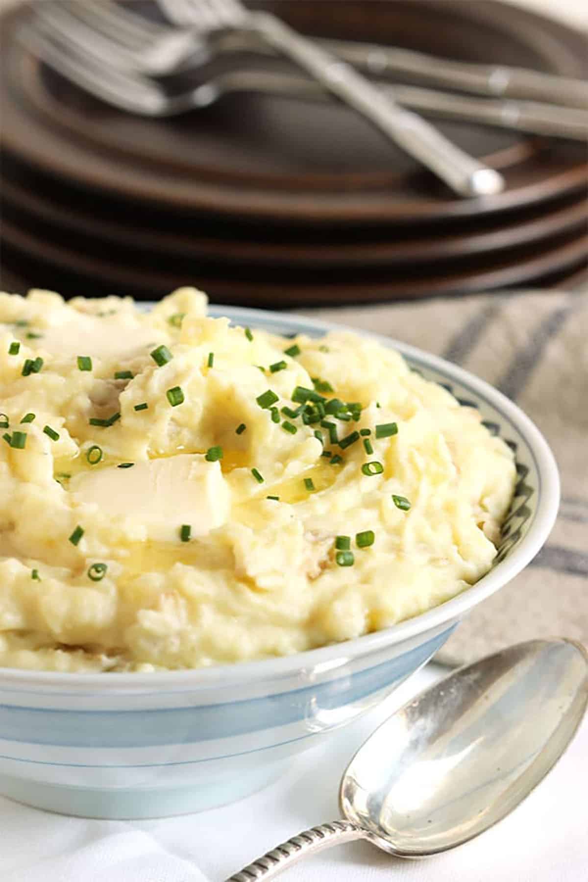 Mashed Potatoes topped with butter and chives in a blue and white stripped bowl with brown plates stacked in the background.
