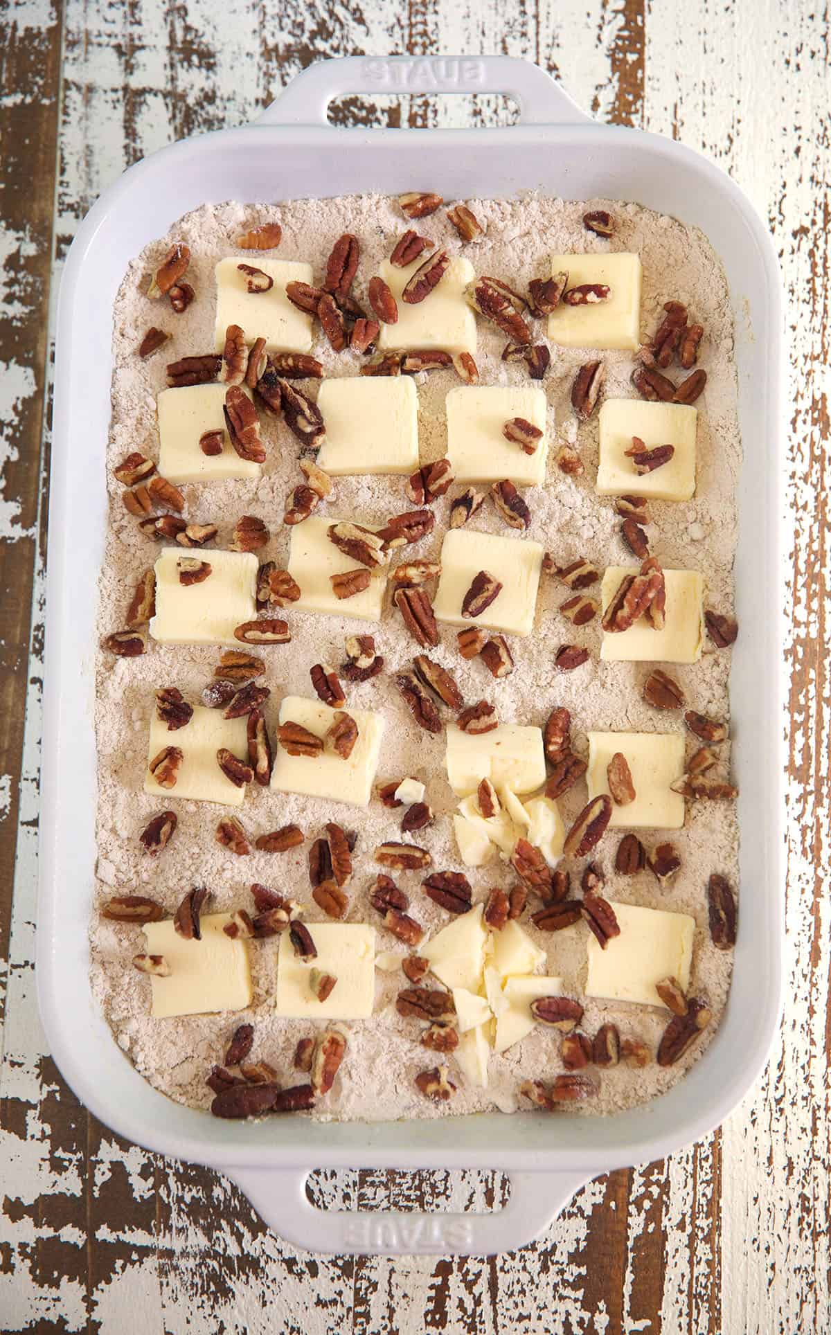 Butter slices and pecans are spread out in a white baking dish on top of cake mix.