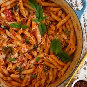 A white and blue dutch oven is filled with cooked penne pomodoro, topped with fresh basil.