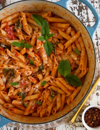 A white and blue dutch oven is filled with cooked penne pomodoro, topped with fresh basil.