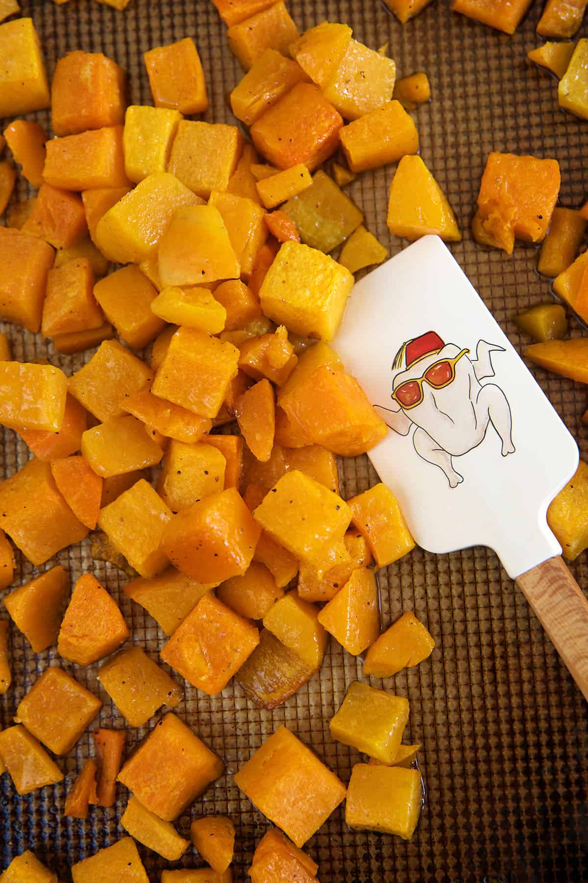 A rubber spatula is mixing chunks of roasted butternut squash on a baking sheet.