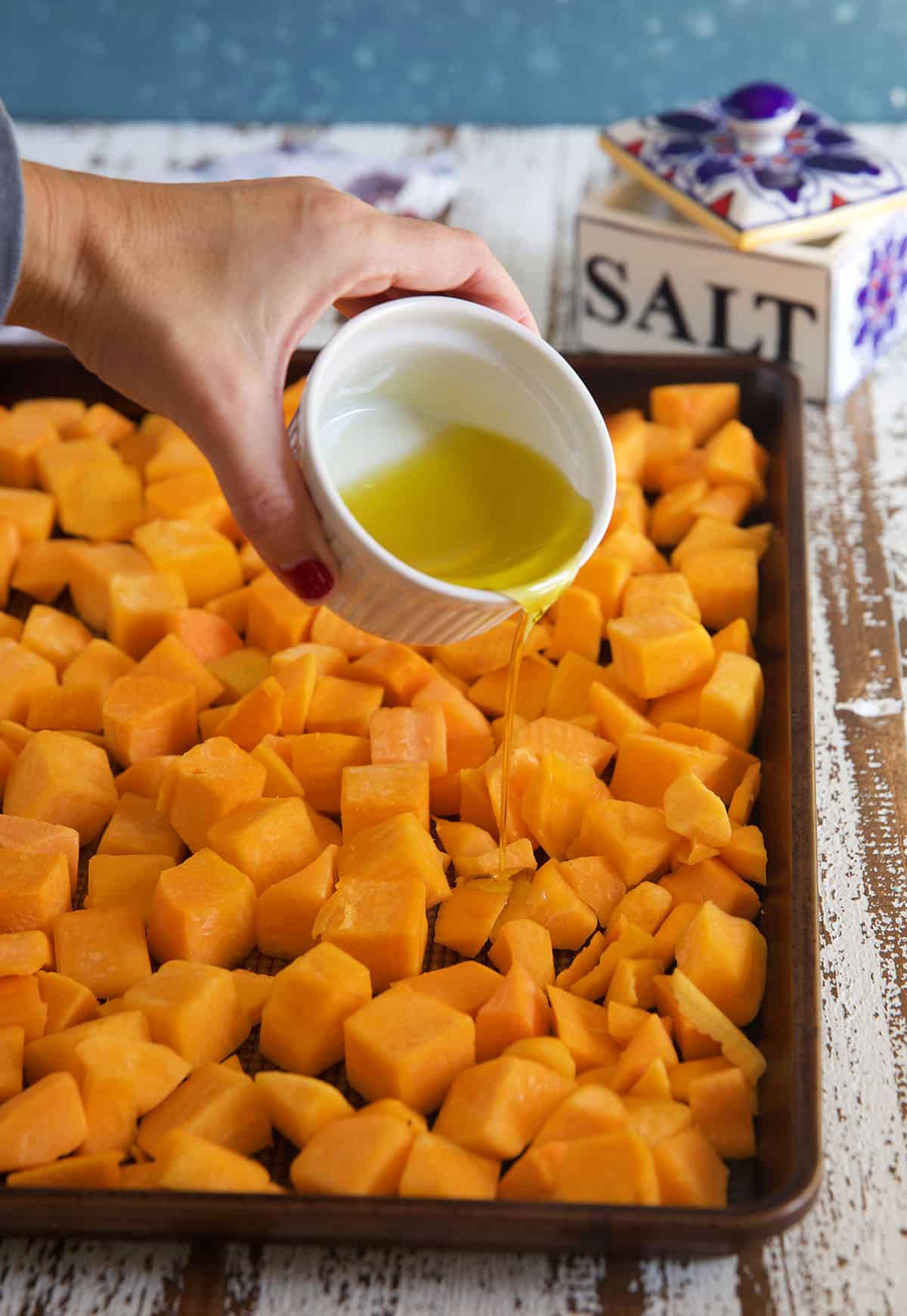 Olive oil is being drizzled over a baking sheet filled with butternut squash.