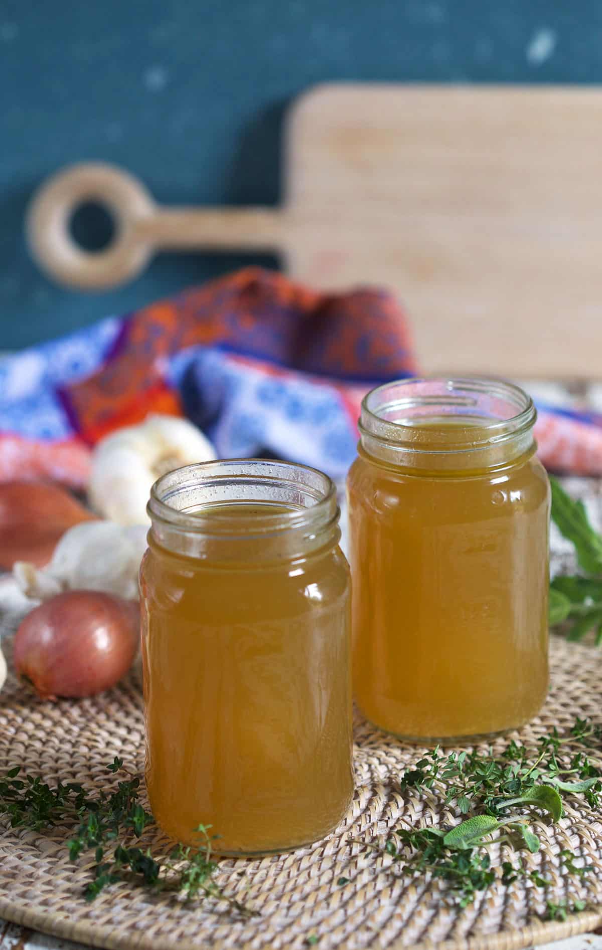 Two full jars of broth are presented on a white surface with a cutting board in the background.