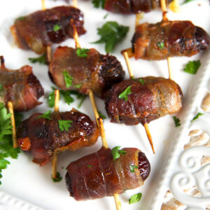 Quite a few stuffed, wrapped dates are presented on a white serving platter.