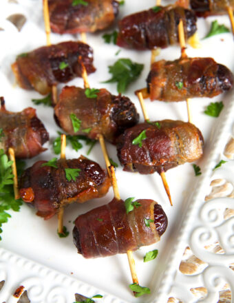 Quite a few stuffed, wrapped dates are presented on a white serving platter.