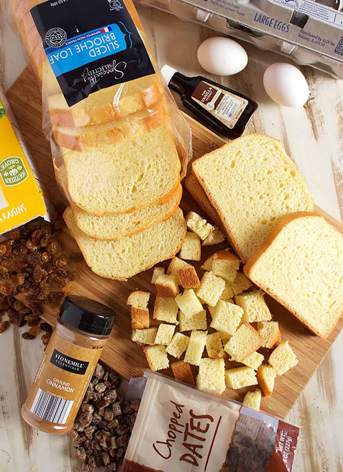 Ingredients for brioche French toast casserole on a board with bread cut into cubes, eggs, cinnamon and nuts.