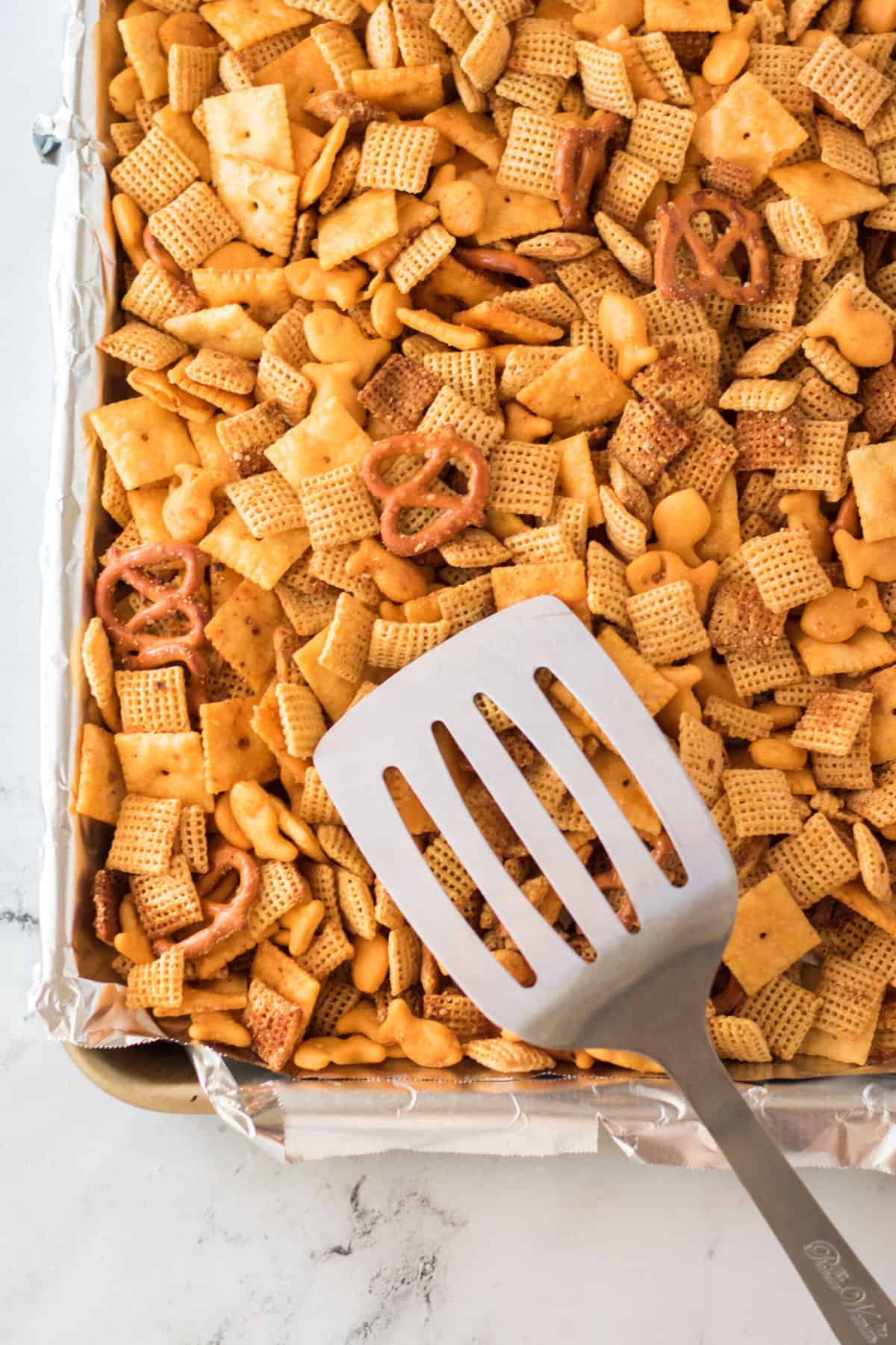 A spatula is hovering over a baking sheet filled with snack mix.