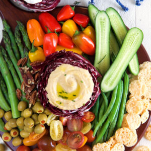 A serving of hummus is surrounded by a rainbow of various veggies on a round platter.