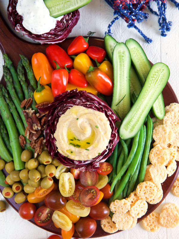 A serving of hummus is surrounded by a rainbow of various veggies on a round platter.