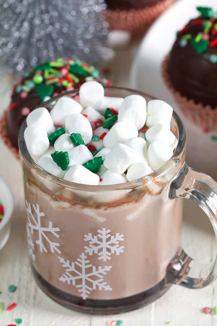 A glass mug is filled with hot cocoa and marshmallows with little green and red sprinkles. 