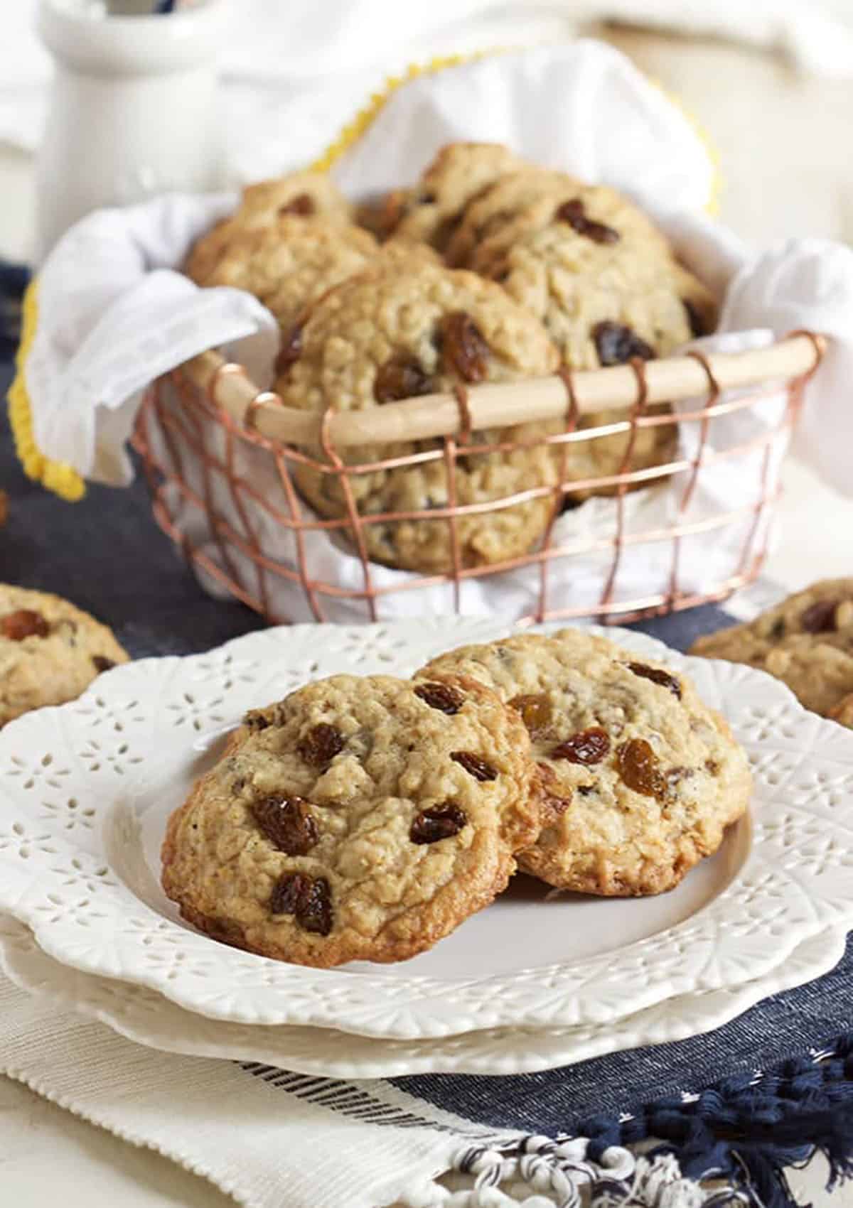 Two oatmeal raisin cookies on a white plate with more cookies in a basket in the background.