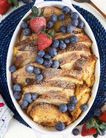 An oval shaped white casserole dish is filled with baked french toast casserole and topped with berries and powdered sugar.