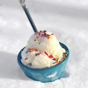Snow ice cream in a blue bowl with a spoon and topped with sprinkles in the snow.