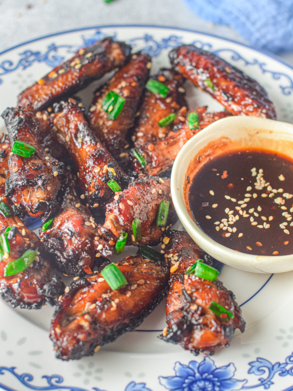 A batch of cooked wings are placed next to a small cup of extra sauce on a blue and white plate.