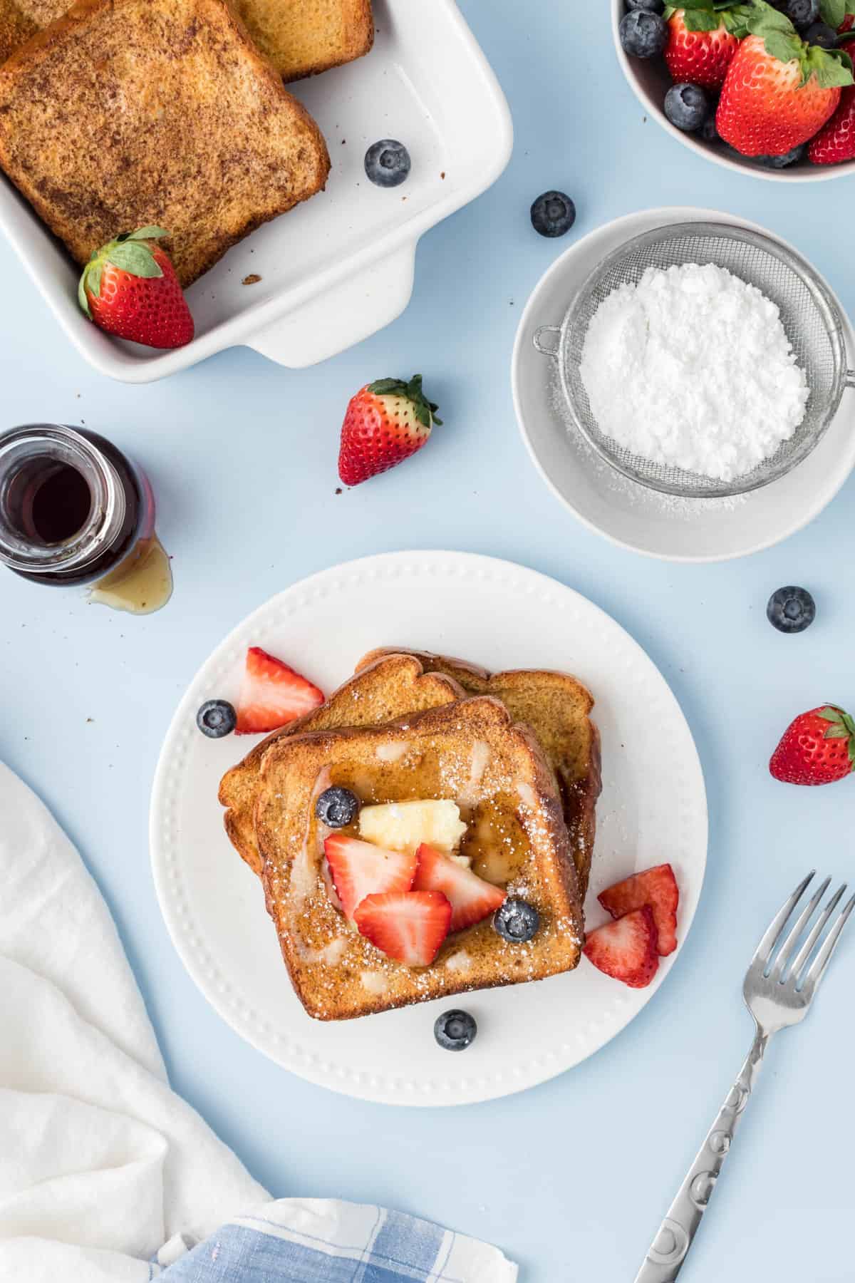 A plate with french toast and berries is placed next to a small bowl of powdered sugar.