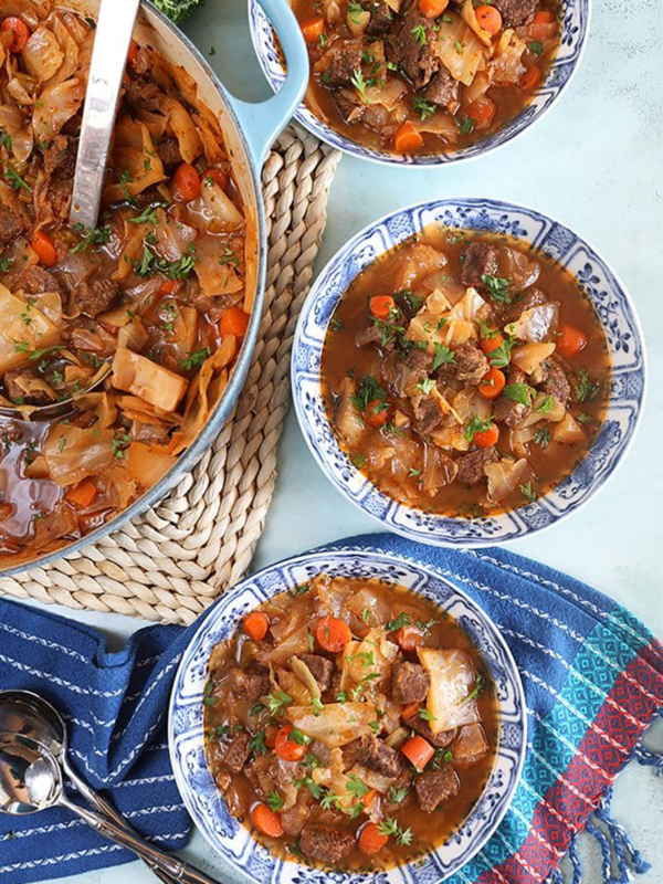 Overhead shot of beef cabbage soup in three blue and white bowls and a pot with a silver ladle.
