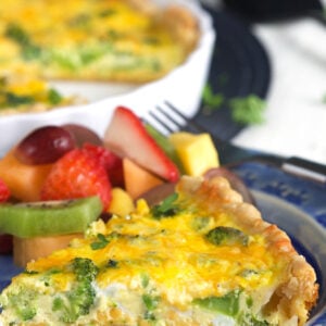 A piece of baked quiche is on a plate next to a fork and fruit.