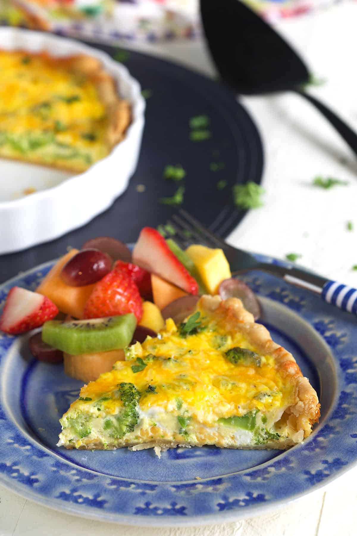 A slice of quiche is presented on a blue and white plate next to a serving of mixed fruit.