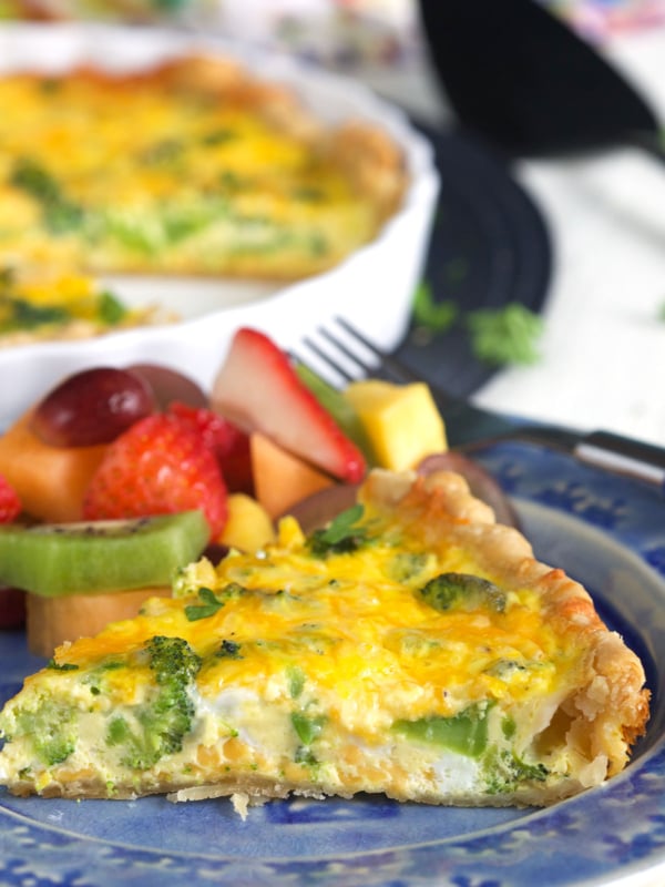 A piece of baked quiche is on a plate next to a fork and fruit.