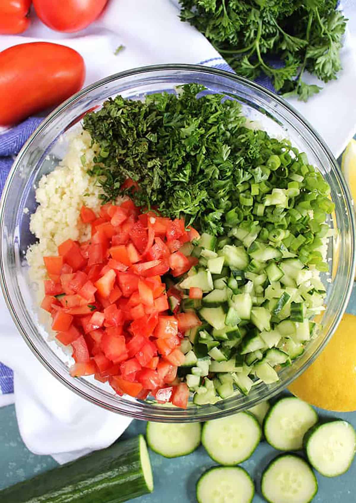 Ingredients for Cauliflower Rice Tabbouleh Salad in a glass bowl with cucumbers, lemons and tomatoes 