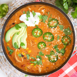 A pot filled with enchilada soup is topped with sliced avocados, jalapenos, and a dollop of sour cream.