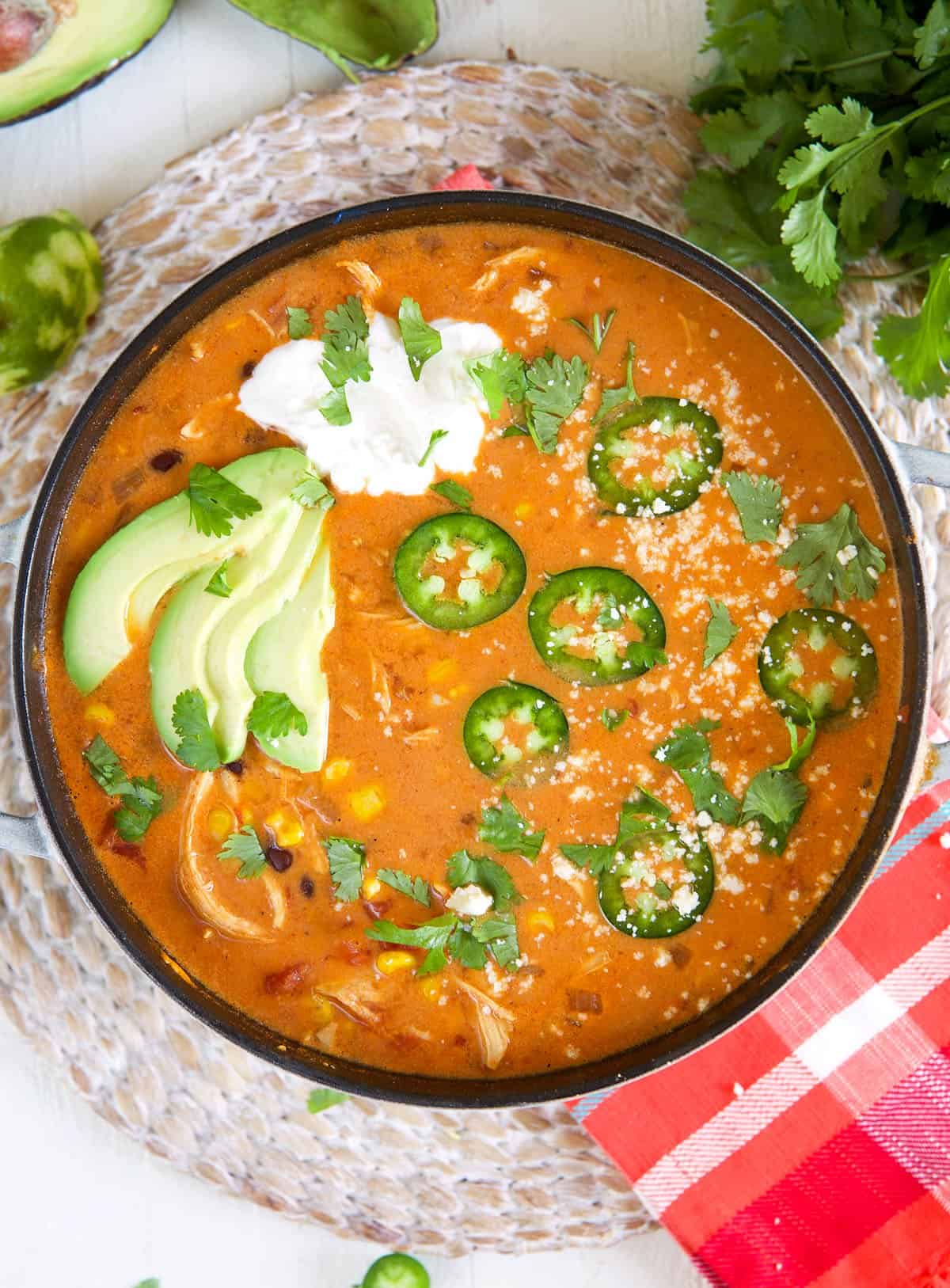 A pot filled with enchilada soup is topped with sliced avocados, jalapenos, and a dollop of sour cream.