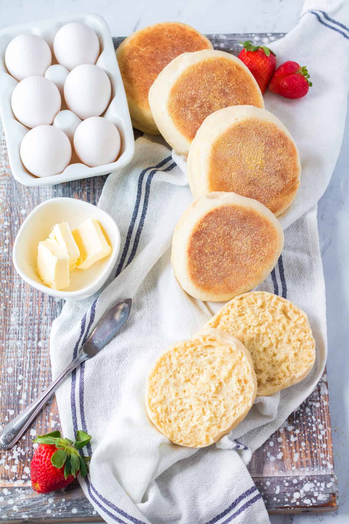 Several english muffins are placed on a countertop next to sliced butter and six eggs.