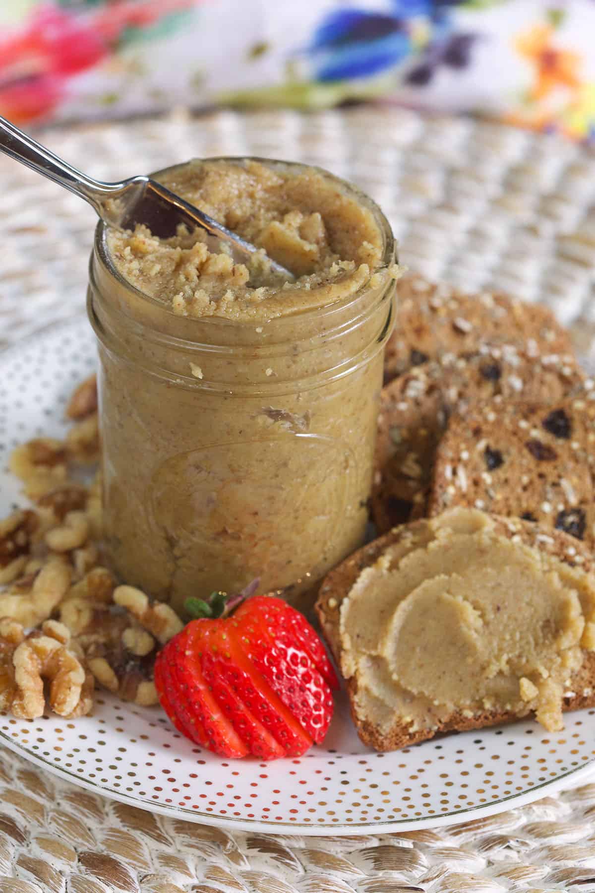 A jar of walnut butter is presented on a white plate next to strawberries and several slices of wheat bread.