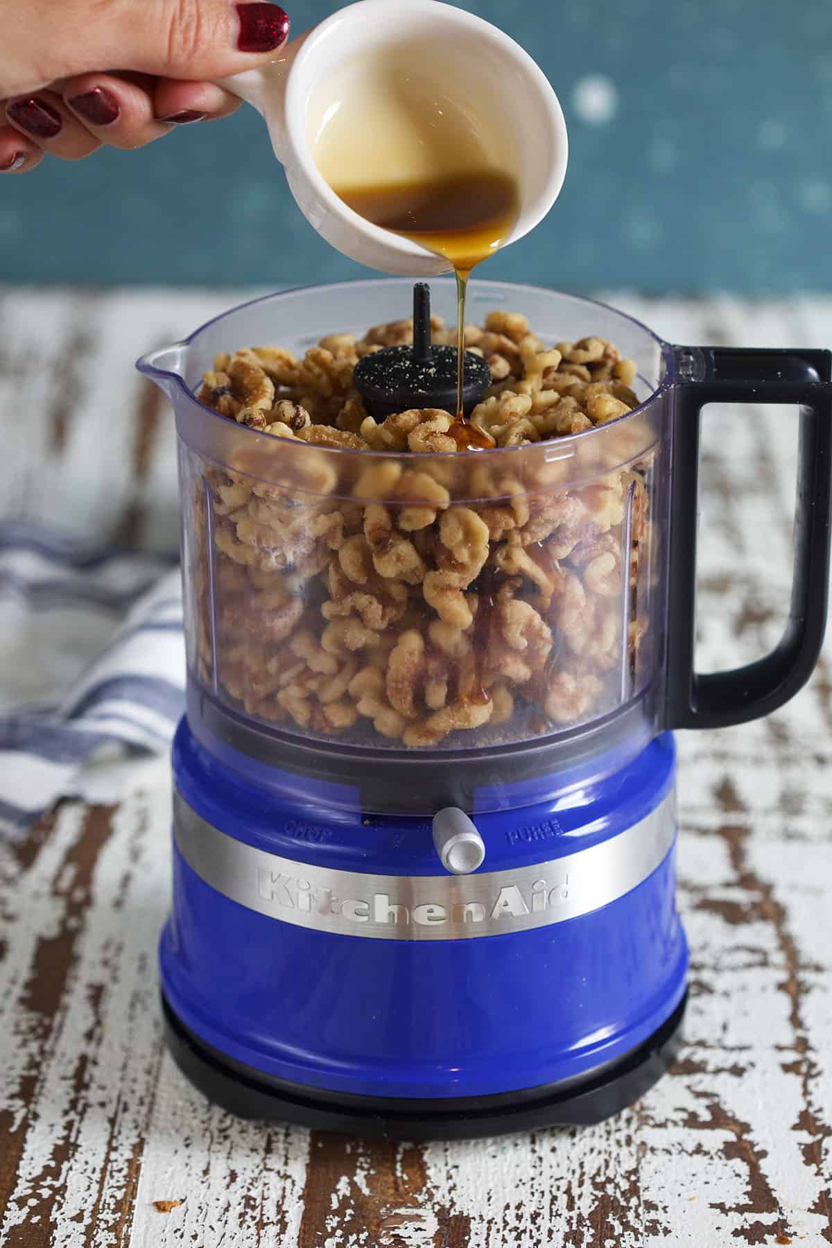 Maple syrup is being poured into a food processor filled with walnuts.