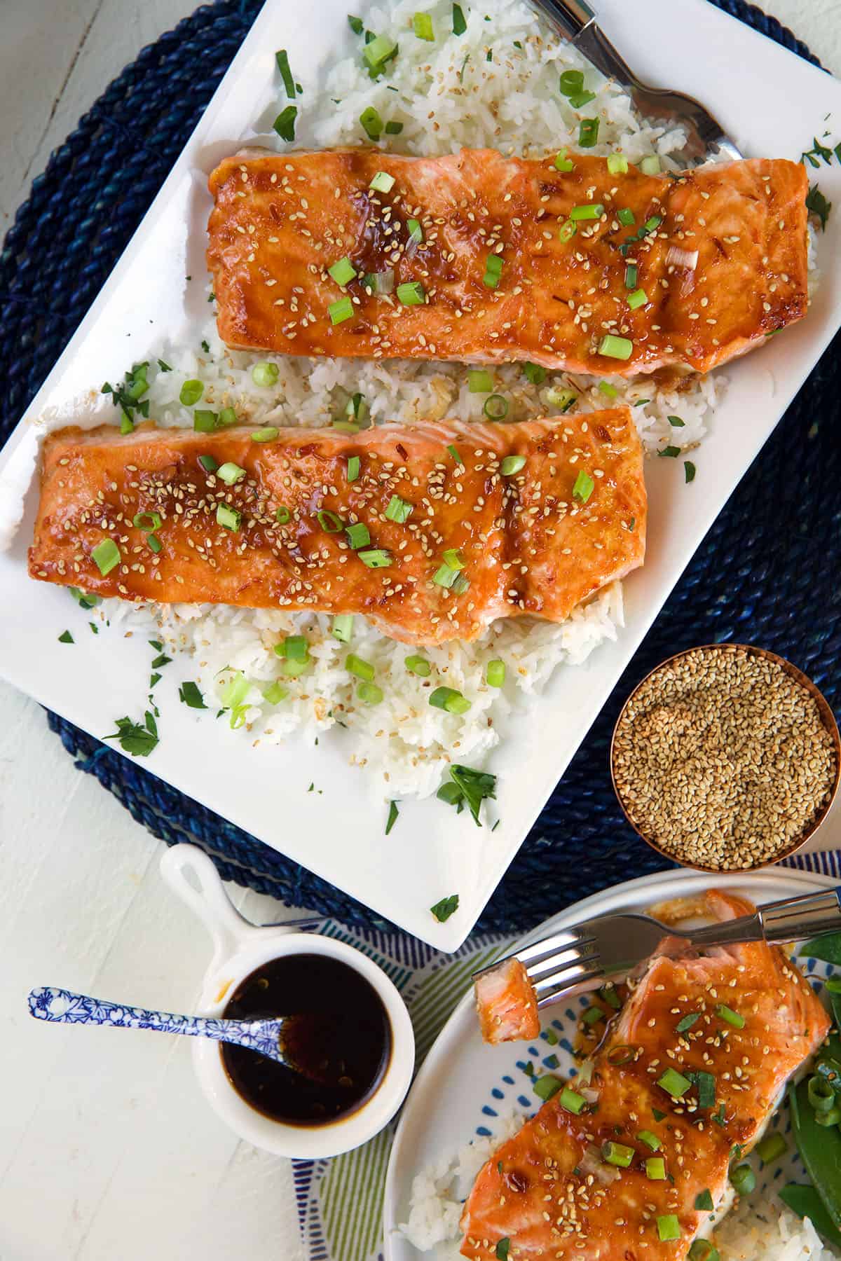 Two cooked salmon filets are placed on a bed of white rice on a rectangular white plate next to a small bowl of brown sauce.