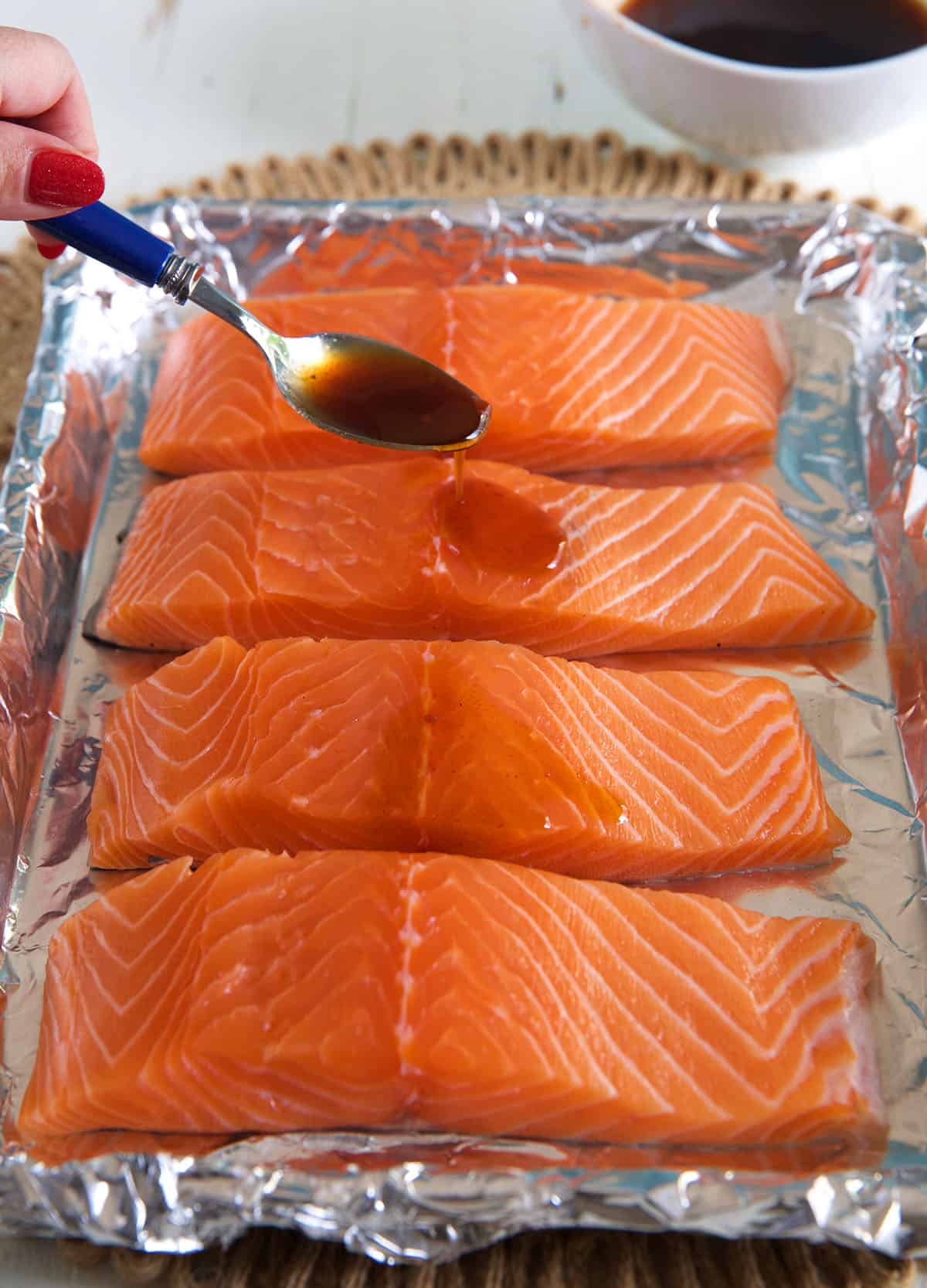 Four salmon filets are on a foil lined baking sheet and one filet is being drizzled with sauce.