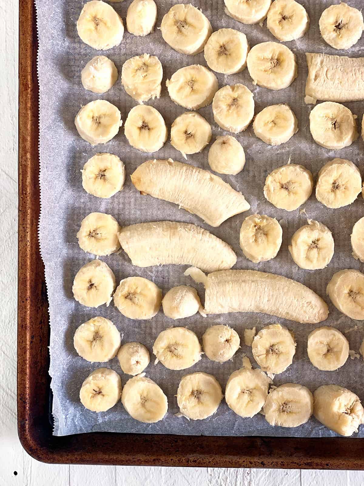 Sliced bananas are spread over a sheet of parchment paper on a baking sheet.