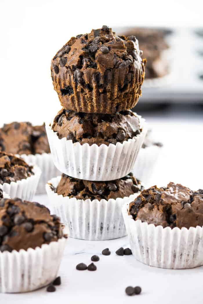 Three chocolate muffins are stacked on top of one another around more muffins on a white surface.