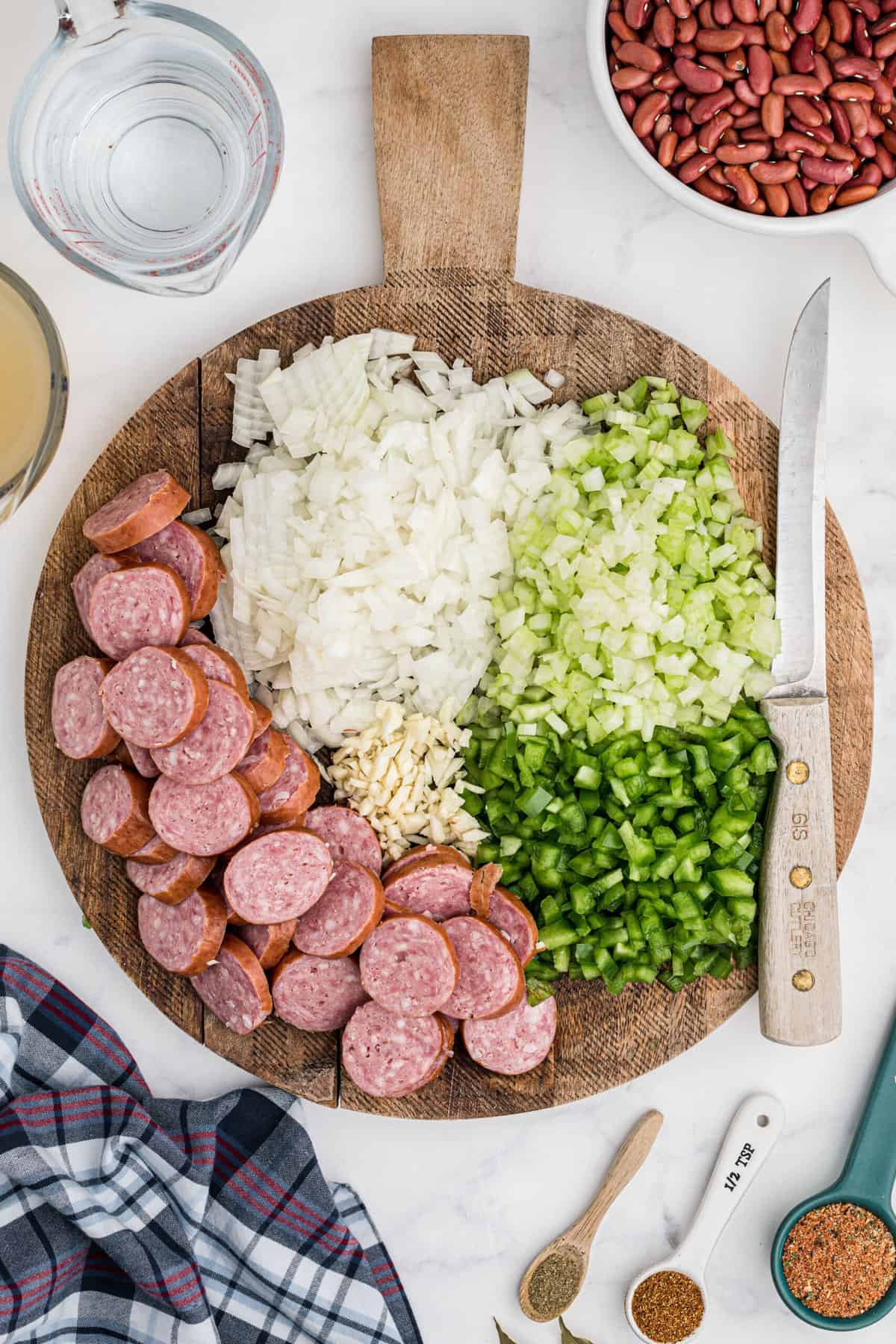 A round wooden cutting board is presented with raw sausage slices and uncooked chopped veggies.