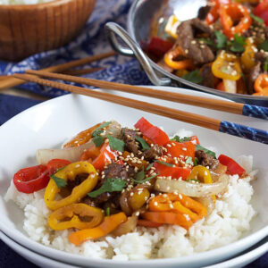 Pepper Steak on white rice in a white shallow bowl with chopsticks resting on top.