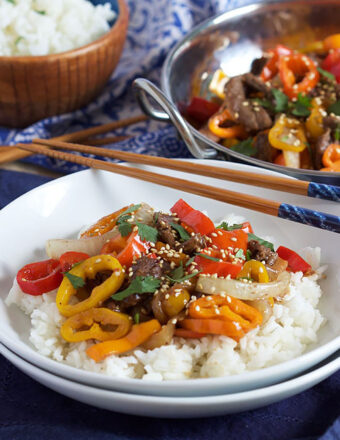 Pepper Steak on white rice in a white shallow bowl with chopsticks resting on top.