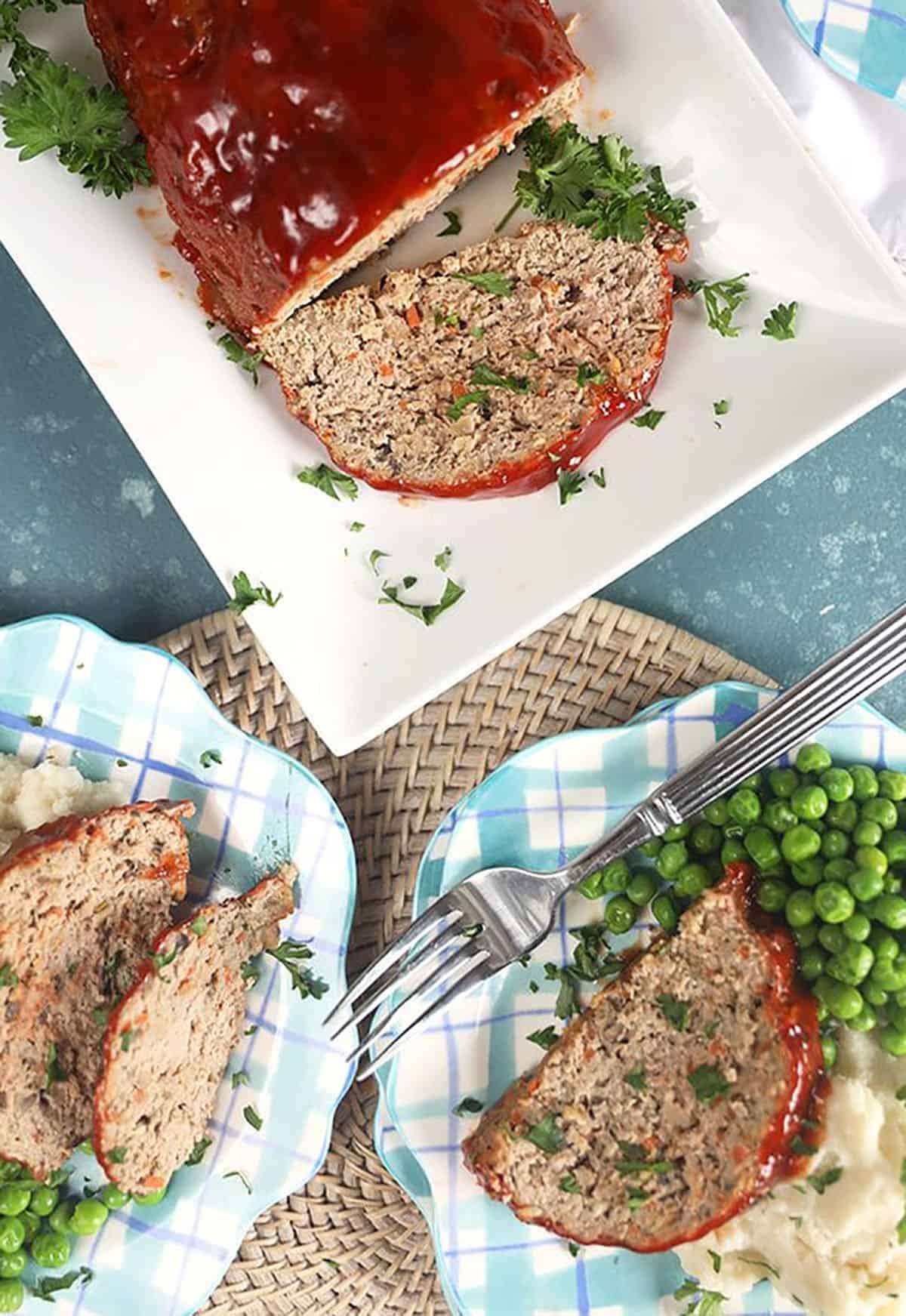 Overhead shot of turkey meatloaf on a white platter with two servings on plaid plates with mashed potatoes and peas.