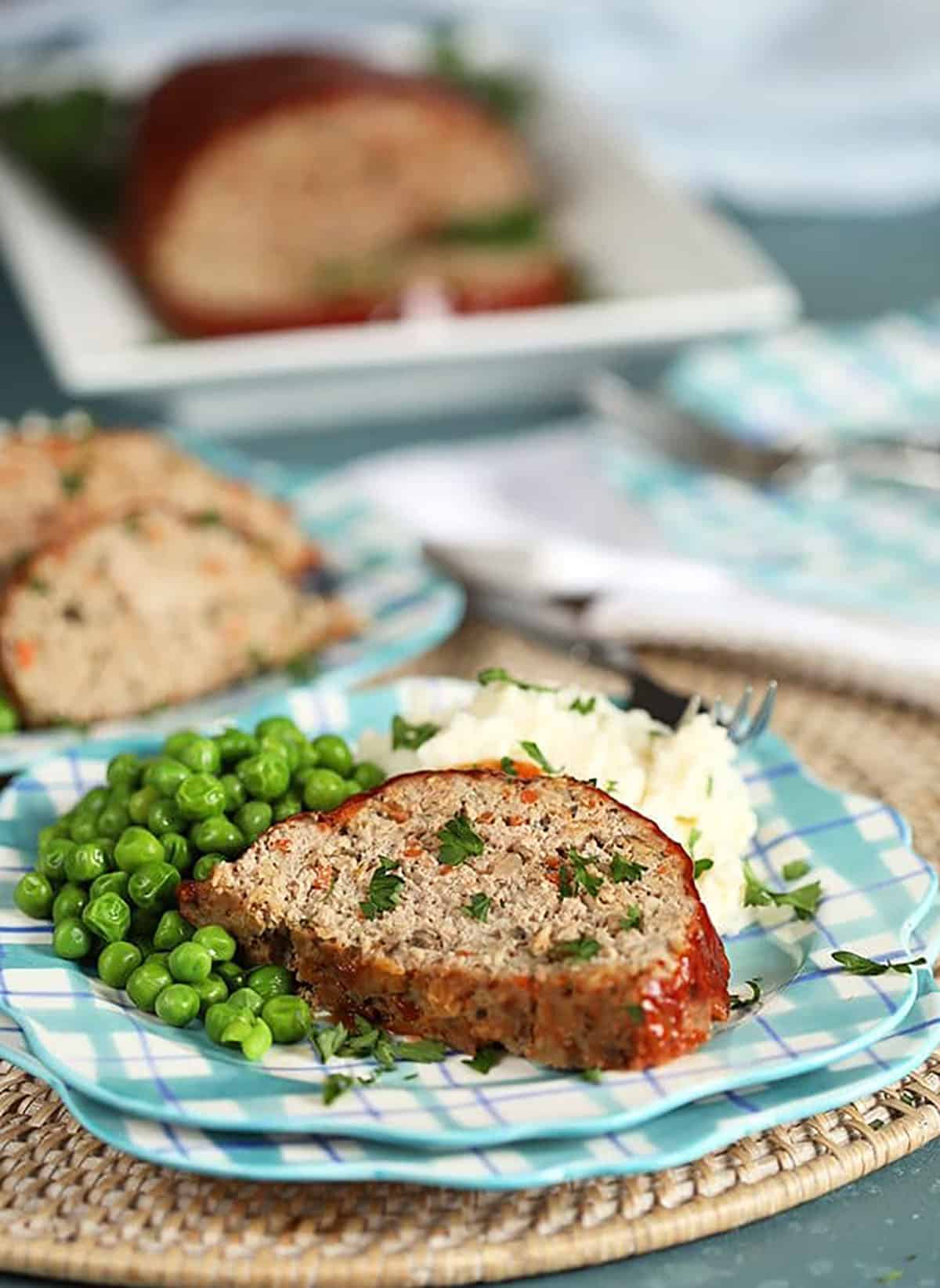 Turkey meatloaf on a plaid plate with mashed potatoes and peas.