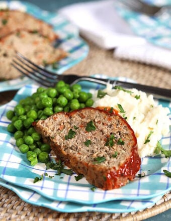 Slice of turkey meatloaf on a blue plaid plate with mashed potatoes and peas.