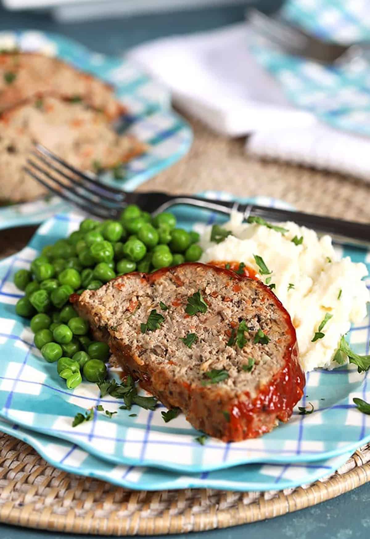 Slice of turkey meatloaf on a blue plaid plate with mashed potatoes and peas.