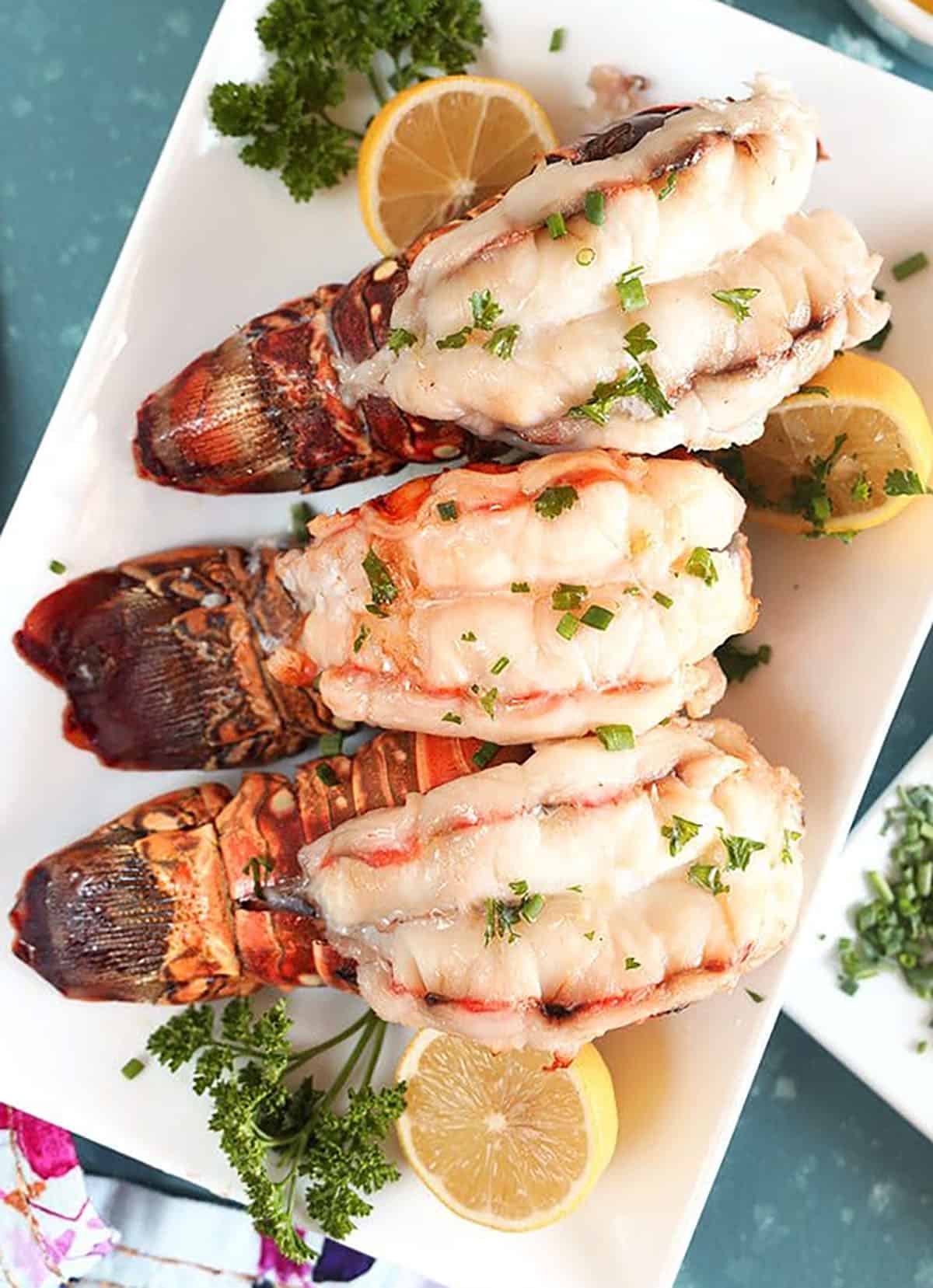 Overhead shot of three baked Lobster Tails with lemons and parsley on a white platter.