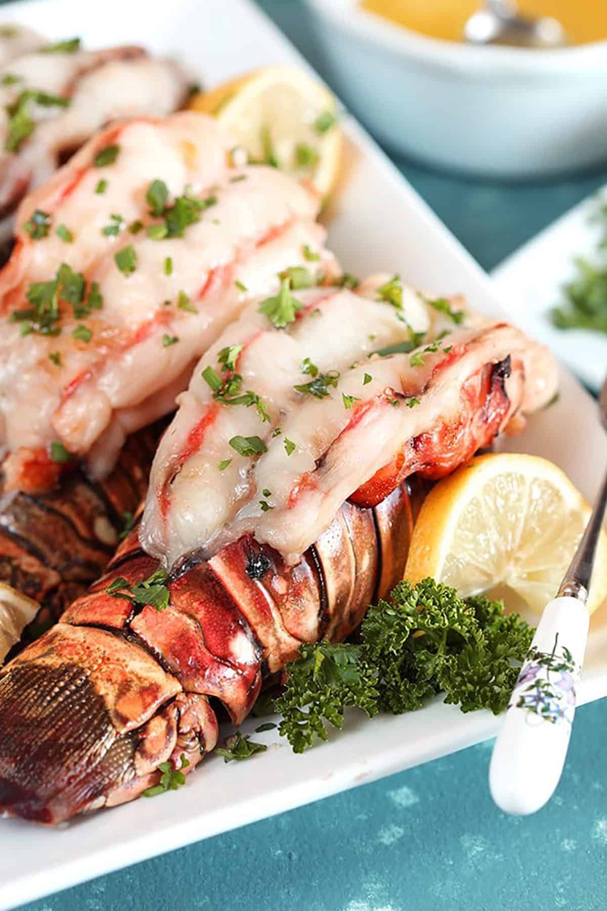Baked lobster tails on a white platter with lemon and parsley.