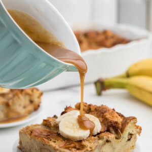 Caramel sauce is being poured over a piece of banana bread pudding on a white plate.