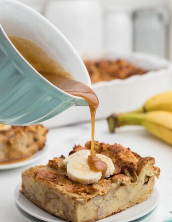 Caramel sauce is being poured over a piece of banana bread pudding on a white plate.